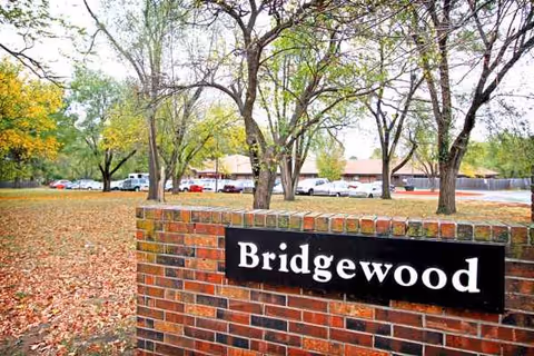 Bridgewood sign mounted on brick wall with trees and parked cars in the background.