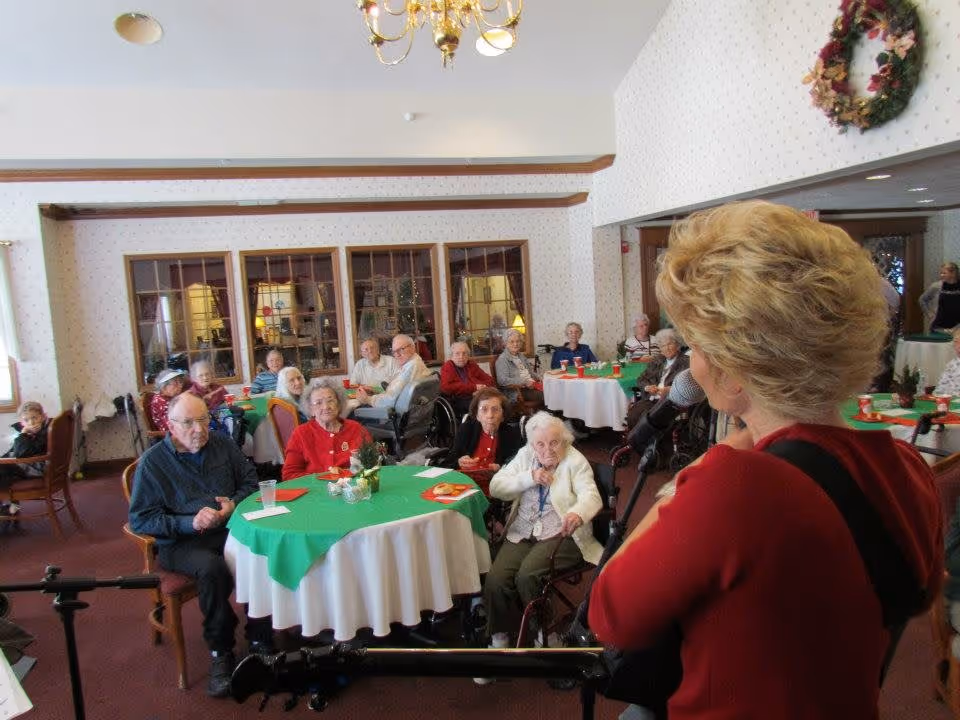 A group of elderly people seated around tables with green and white tablecloths in a decorated room, attentively listening to a woman with short blonde hair speaking or singing into a microphone. The room has a chandelier, wreath decorations, and large windows with wooden frames.