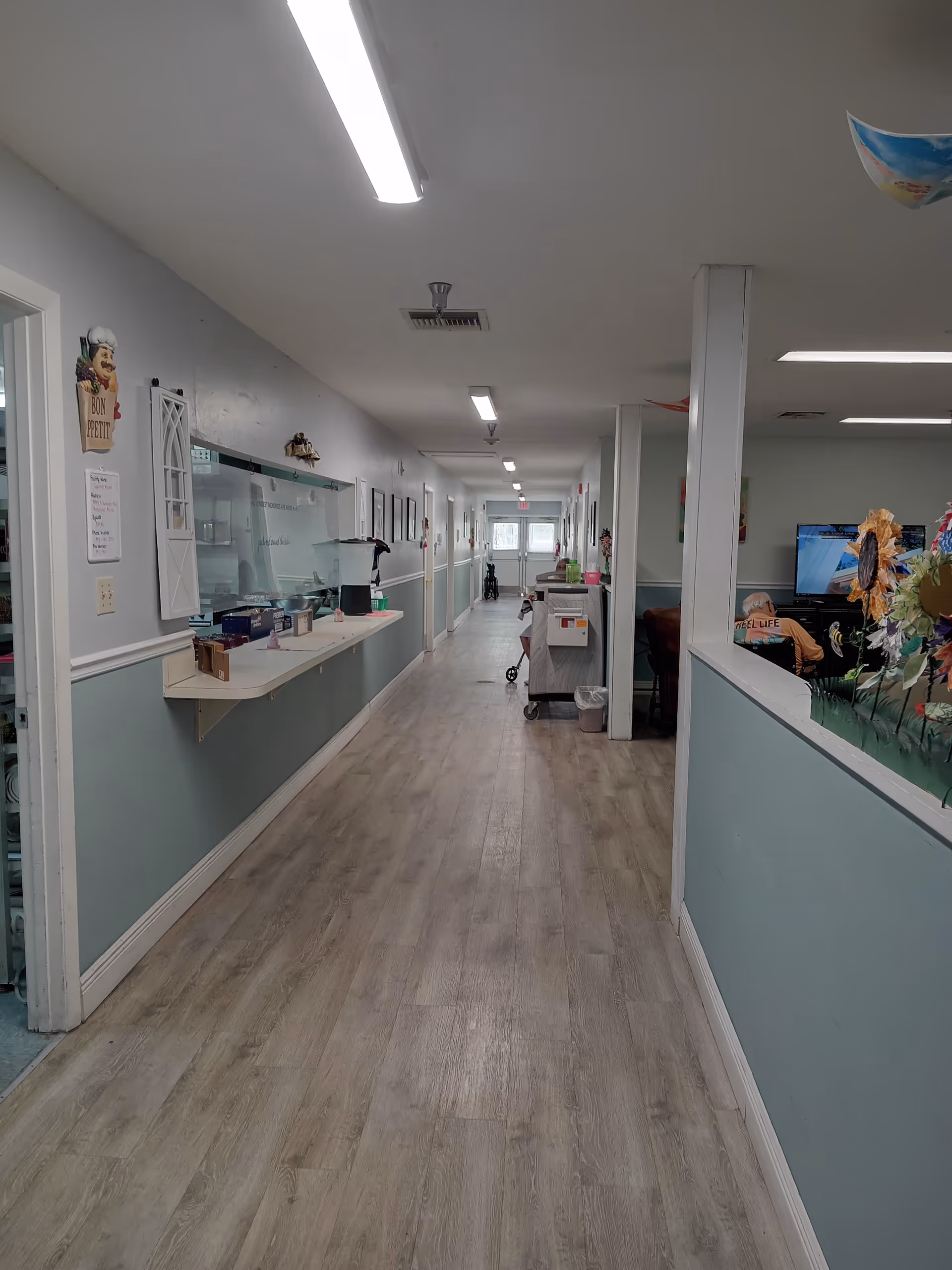 A long hallway inside Sugarmill Manor Assisted Living with light wood flooring and light blue and white walls. On the left side, there is a counter with various items and a window opening to another room. The hallway is well-lit with ceiling lights and has doors and framed pictures along the walls. On the right side, there is a partial view of a common area with a television and decorative flowers.