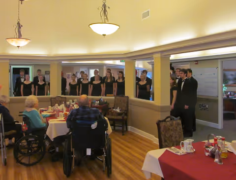Residents seated at dining tables watch a formally dressed youth choir performing along a half wall in a communal dining room.