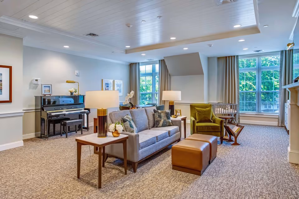 A bright and spacious living room area with a gray sofa, green armchair, two brown ottomans, and two side tables with lamps. There is a black upright piano against the wall with framed pictures above it. Large windows with beige curtains allow natural light to fill the room, and a fireplace is visible on the right side.