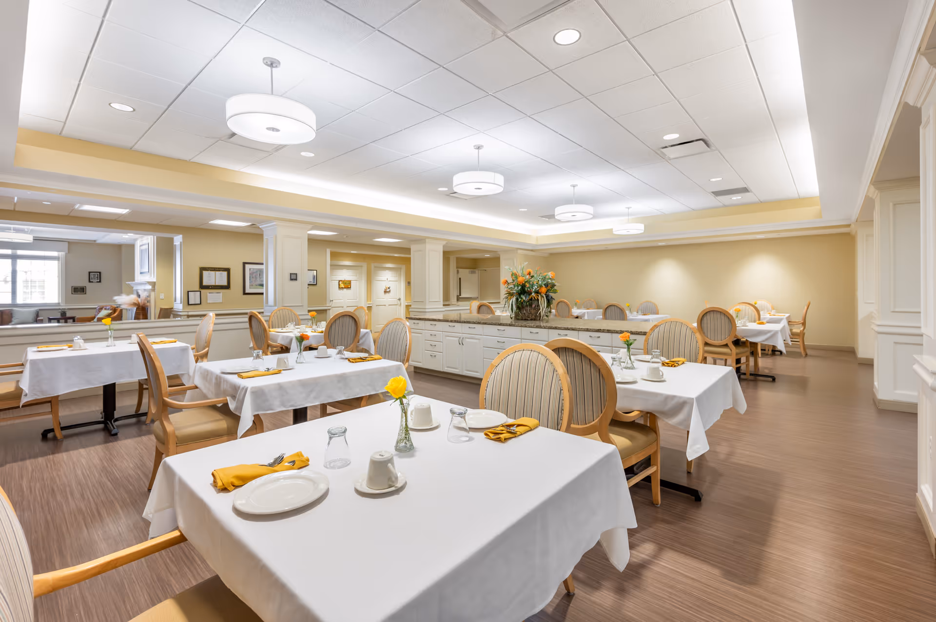 A bright and spacious dining room with several tables covered in white tablecloths, each set with plates, cups, glasses, and yellow napkins. The room features wooden chairs with cushioned seats and backs, a central island with a flower arrangement, and soft yellow walls. Ceiling lights provide ample illumination, and a seating area with a fireplace is visible in the background.