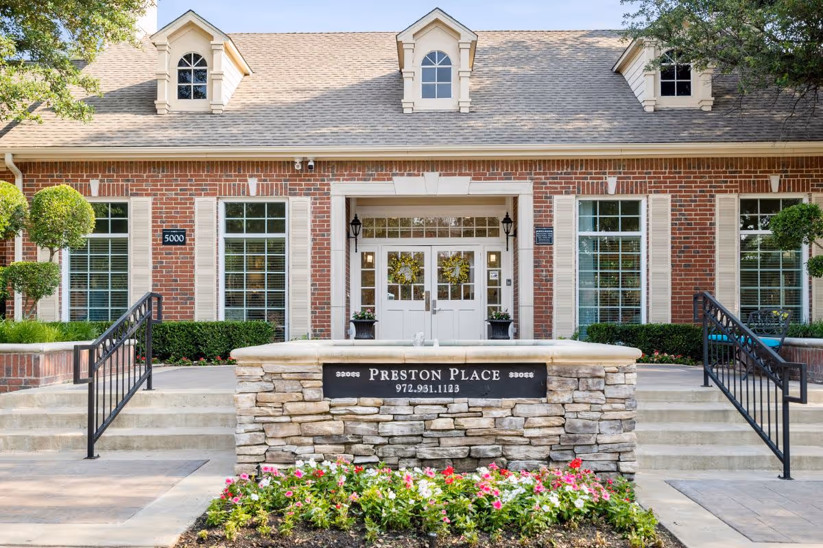 Front entrance of a brick building marked "Preston Place" with a stone sign, steps, double doors, and a flowerbed.