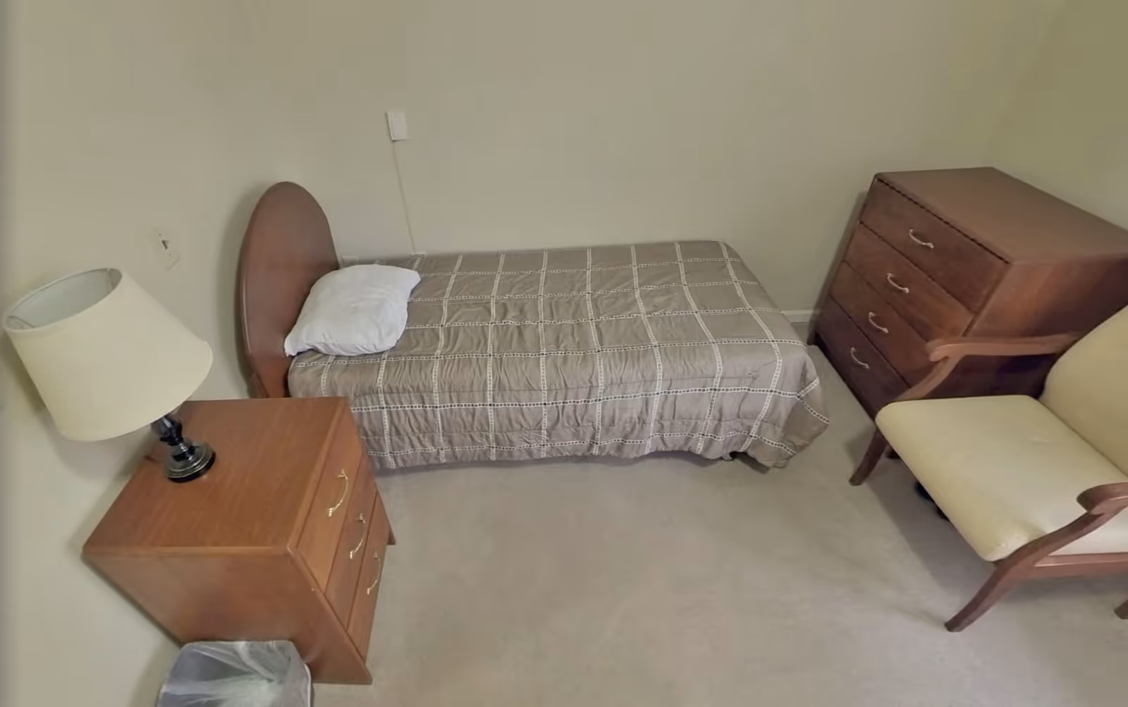 A small bedroom with a single bed covered with a beige checkered bedspread and a white pillow. Next to the bed is a wooden nightstand with a lamp on it. Across from the bed is a wooden dresser with four drawers and a beige upholstered armchair with wooden armrests. The room has light-colored walls and carpeted floor.