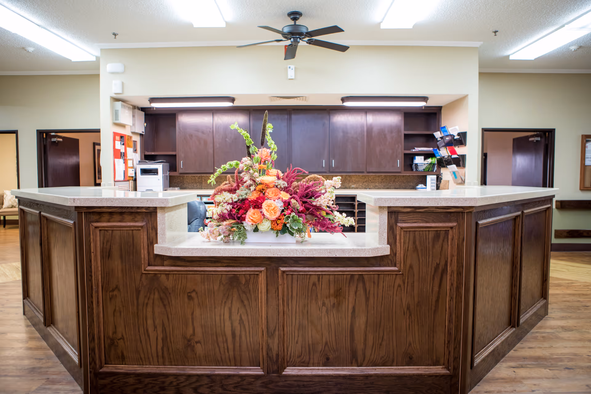 Reception desk area in a nursing and rehabilitation center with a large floral arrangement on the counter, dark wood cabinetry behind the desk, and a ceiling fan above.