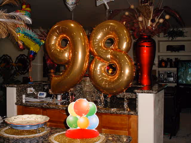 Indoor scene showing large gold balloons shaped as the numbers 9 and 8 placed on a kitchen counter with granite countertop. There are colorful balloon decorations and a large red vase in the background, along with kitchen cabinets and appliances.