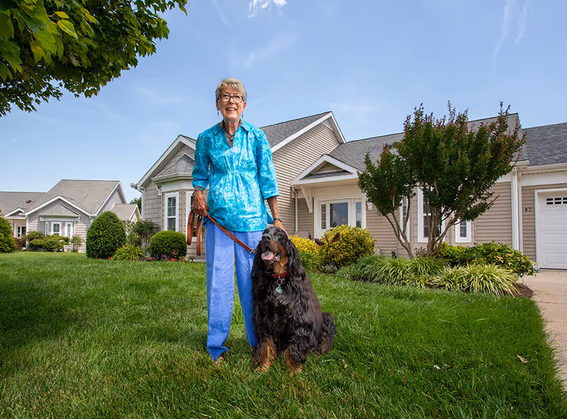 An elderly woman wearing a blue shirt and light blue pants stands on a green lawn holding a leash attached to a large black and brown dog. Behind them are beige single-story houses with well-maintained bushes and trees under a clear blue sky.