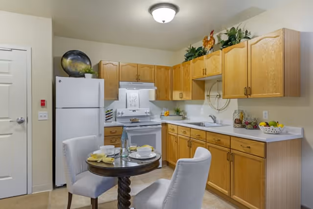 A kitchen area with wooden cabinets, a white refrigerator, stove, and sink. A small round dining table with two upholstered chairs is set with plates, cups, and utensils. Decorative plants and items are placed on top of the cabinets and countertop.
