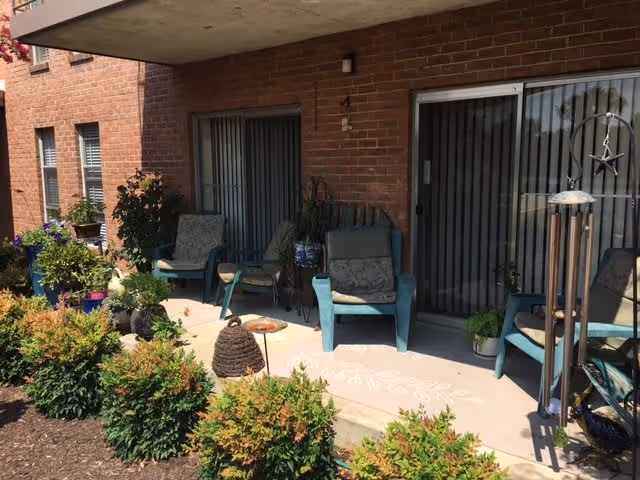 Outdoor patio area of Brooklawn Apartments with several cushioned chairs, potted plants, a wind chime, and decorative items on a concrete porch in front of a brick building with sliding glass doors and windows.