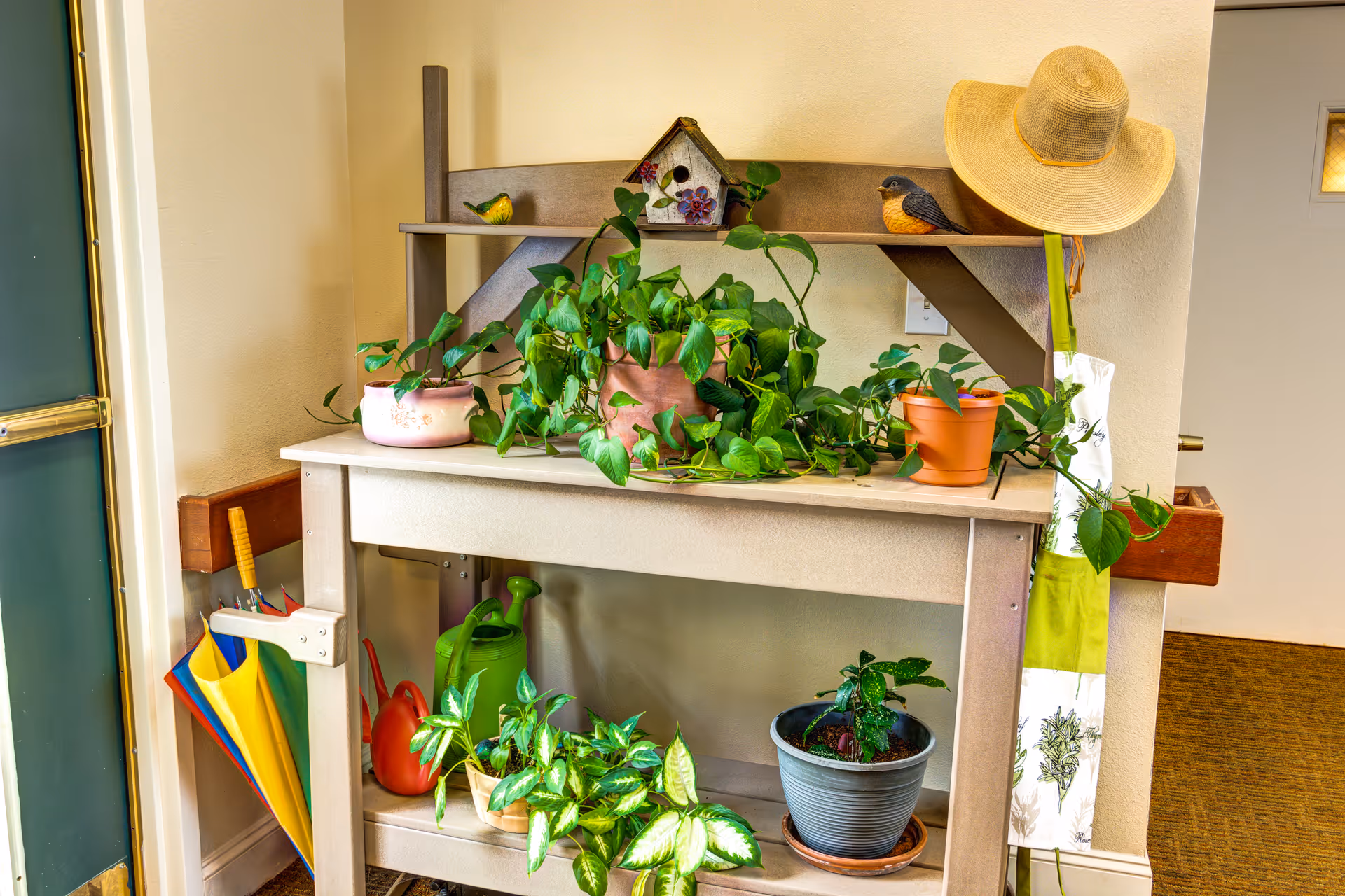 A small wooden gardening table against a beige wall, decorated with several potted green plants, a birdhouse, two bird figurines, a straw hat hanging on the side, and colorful umbrellas stored in a holder attached to the table. There is a green watering can and other gardening items on the lower shelf.
