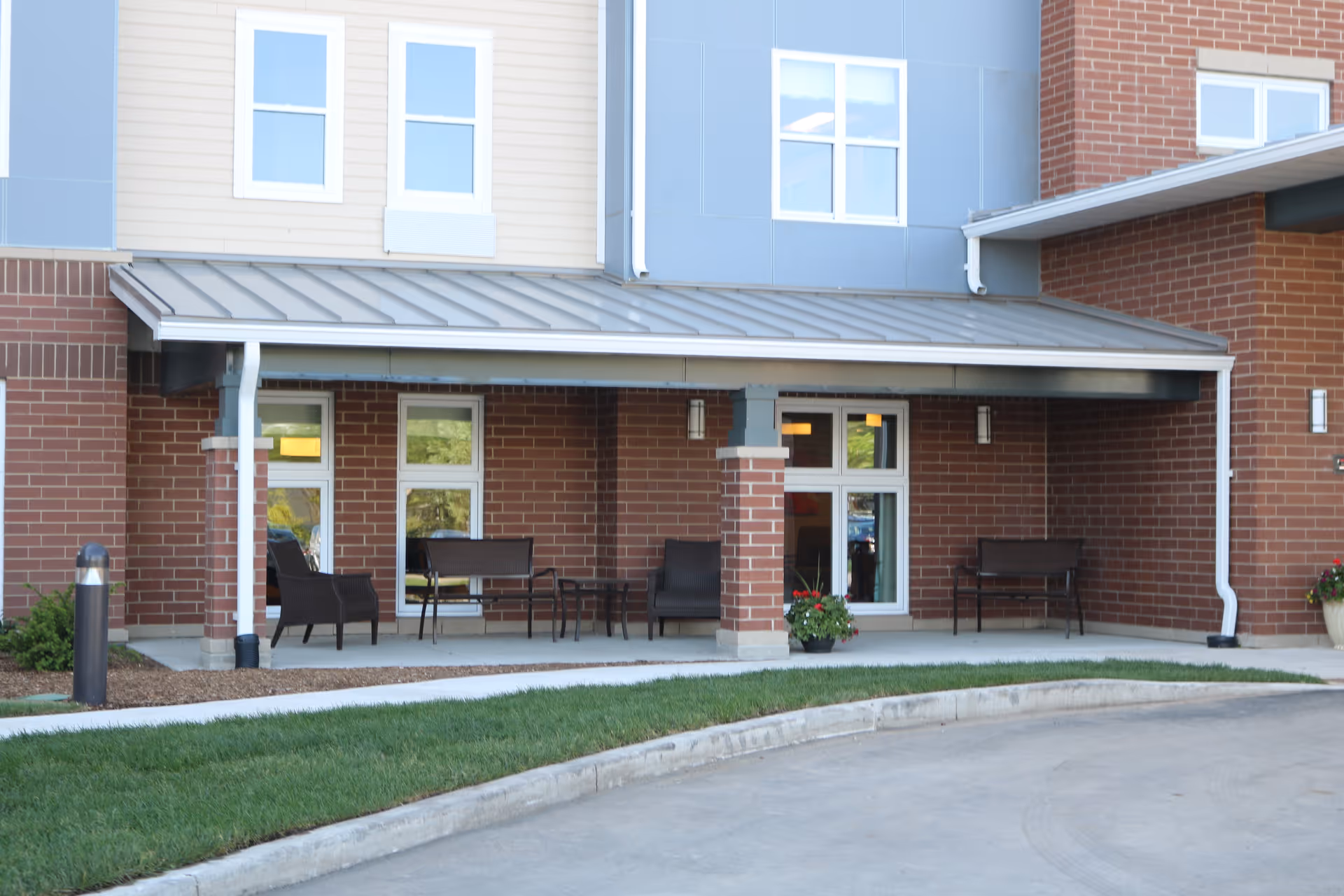 Covered outdoor seating area at the entrance of a brick and siding building with several chairs, benches, potted plants, and windows.