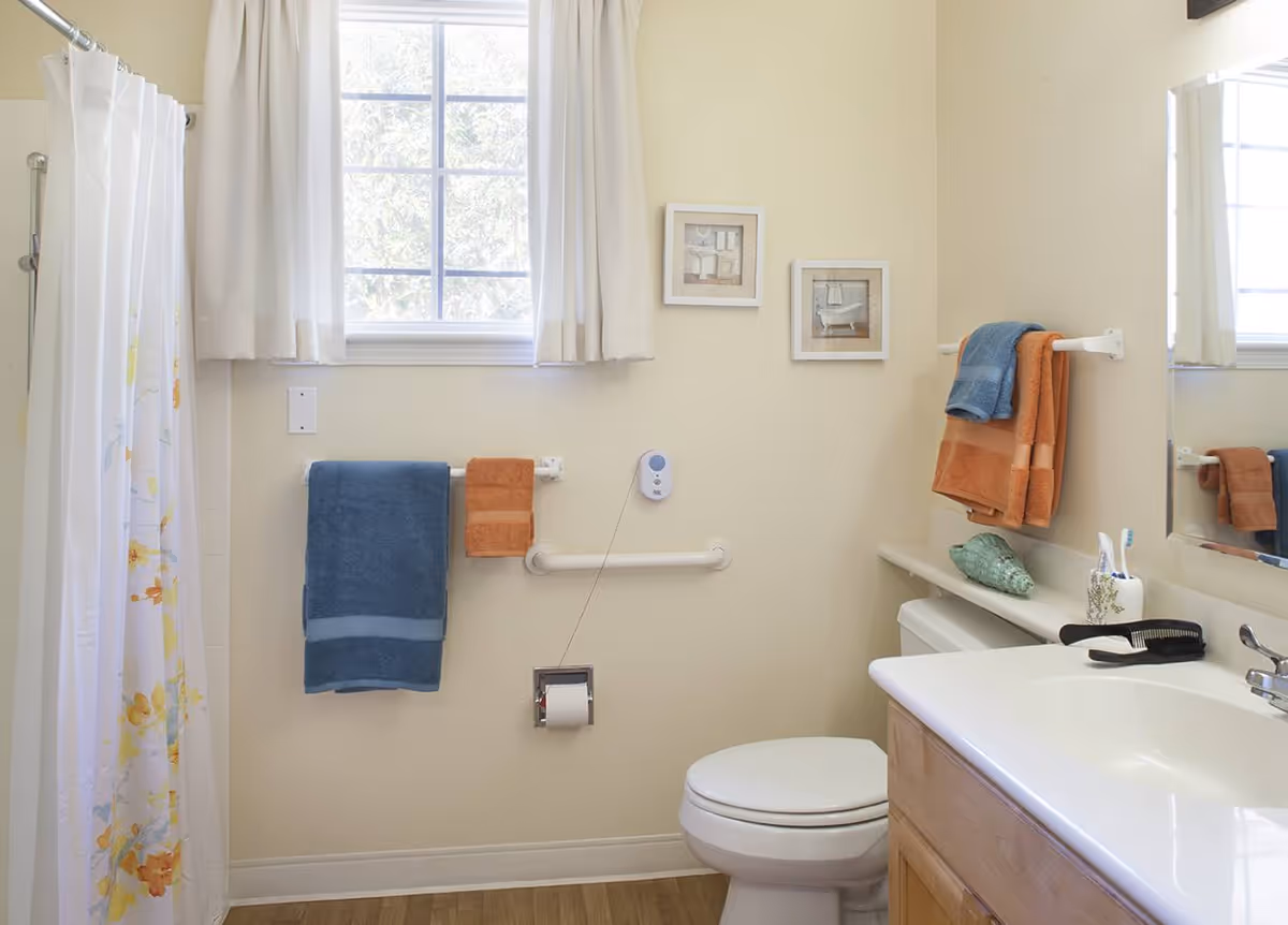 Bright accessible bathroom showing a shower with curtain, toilet, sink, grab bar, and colorful towels beneath a window.