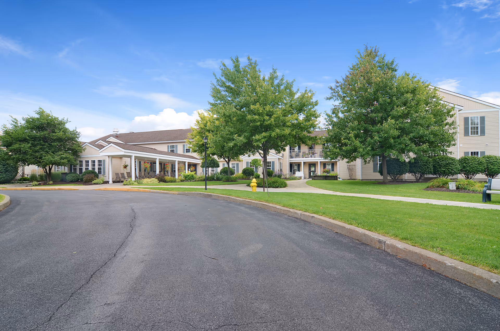 Exterior view of Brookdale Summerfield senior living facility showing a large, two-story beige building with white trim, surrounded by green trees and well-maintained lawns under a blue sky with some clouds. A curved paved driveway leads to the entrance with a covered porch area featuring seating.