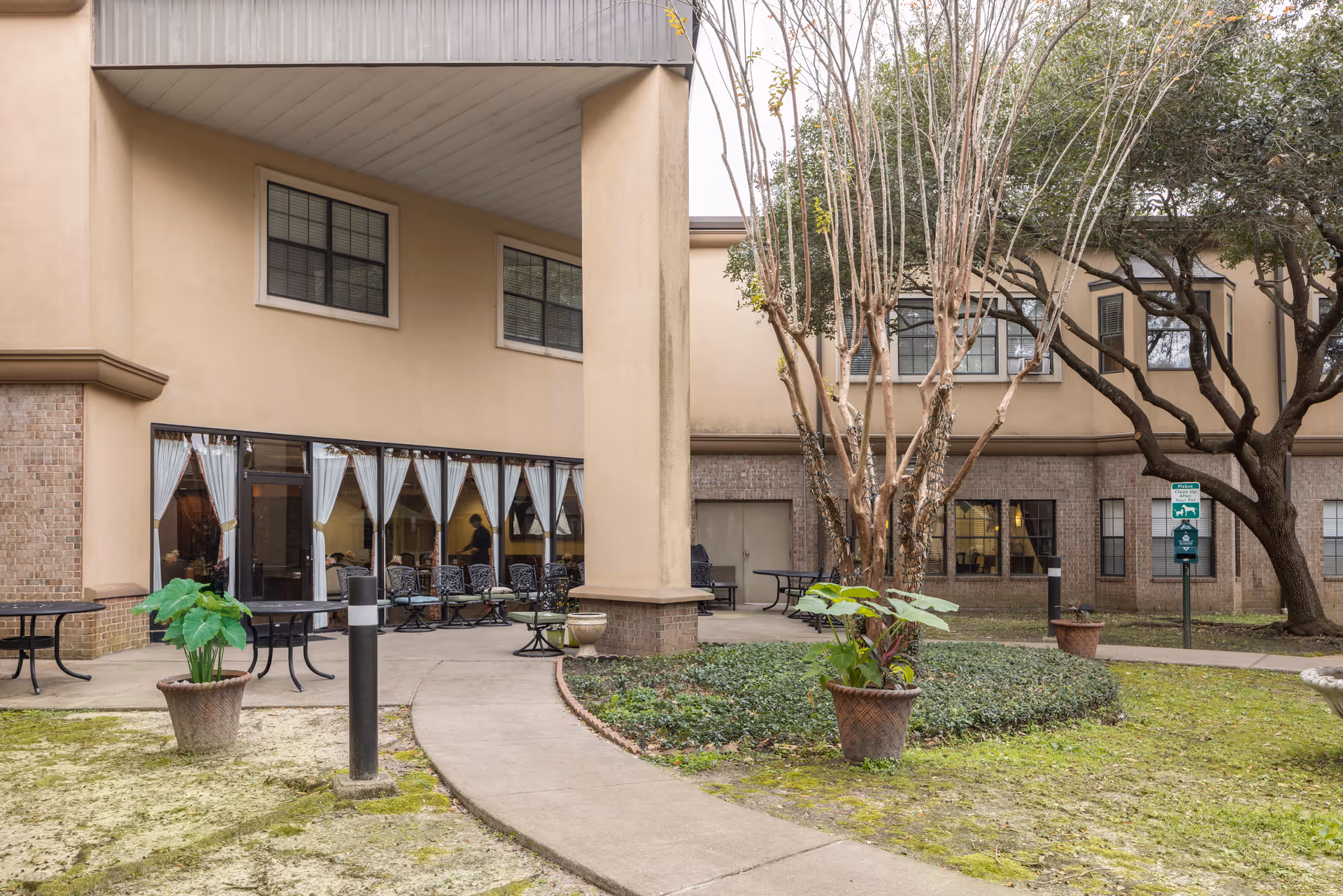 Outdoor courtyard area of a senior living facility with a paved walkway, potted plants, trees, and patio tables and chairs near large windows with white curtains.