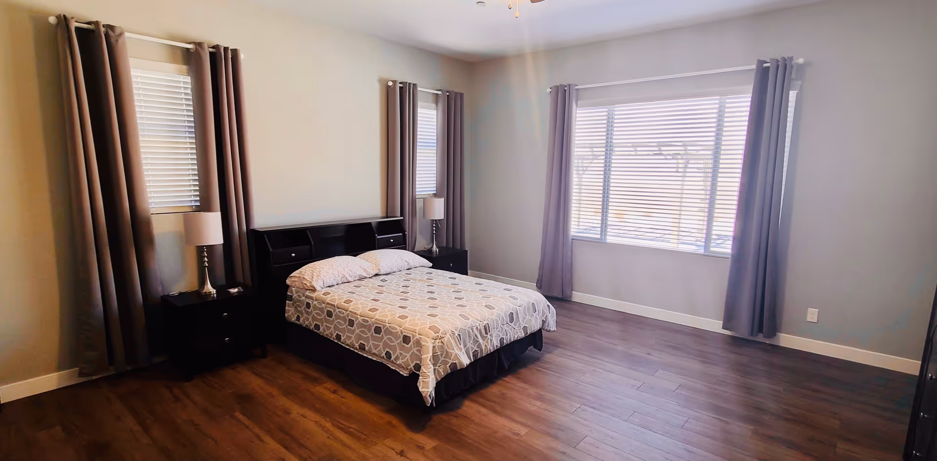 A spacious bedroom with a double bed covered in a patterned bedspread, two bedside tables each with a lamp, three windows with blinds and gray curtains, and wooden flooring.