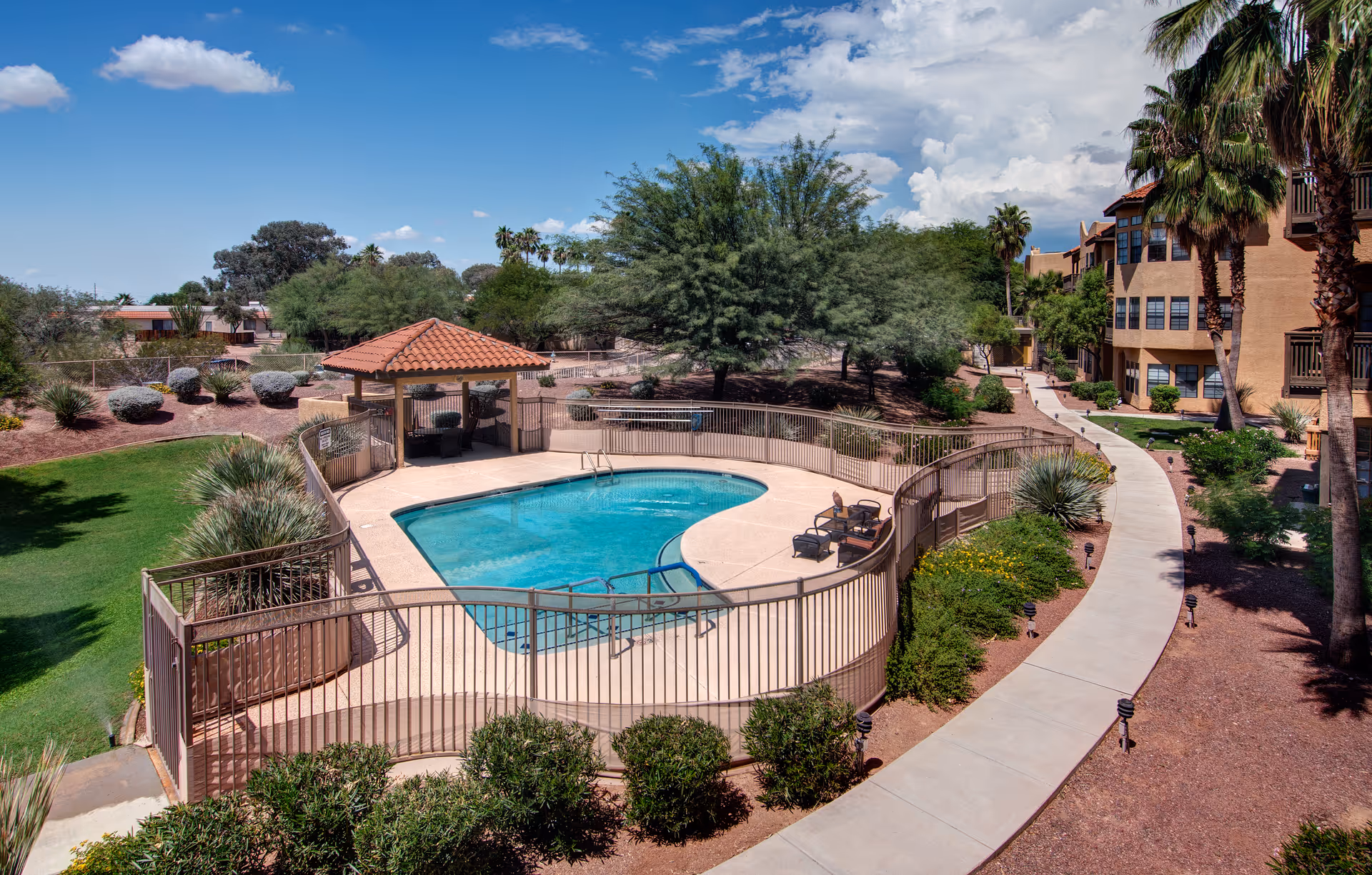 Outdoor swimming pool area surrounded by a safety fence with a small covered seating area nearby. The pool is located next to a curved walkway and landscaped garden with bushes, palm trees, and other greenery. A multi-story building is visible on the right side under a partly cloudy blue sky.