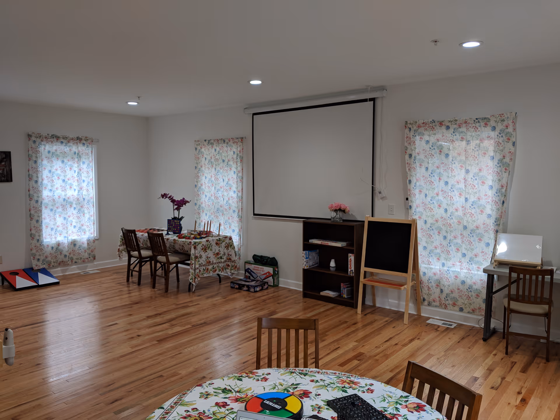 A spacious room with wooden flooring and white walls featuring three windows covered with floral curtains. There are two tables with floral tablecloths, each surrounded by wooden chairs. One table has a vase with purple flowers, and the other table has a board game on it. A small bookshelf with board games and a vase with pink flowers is against the wall, next to a blackboard on a wooden stand. A projector screen is mounted on the wall above the bookshelf. The room is well-lit with ceiling lights.