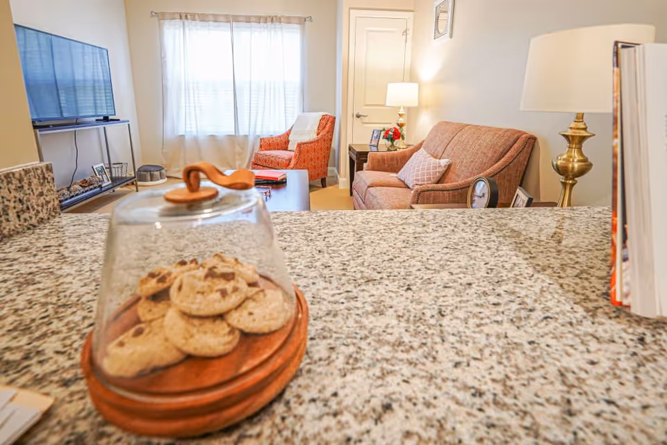 View from a kitchen counter showing cookies under a glass dome in the foreground and a cozy living room with a sofa, armchair, TV, and lamps in the background.