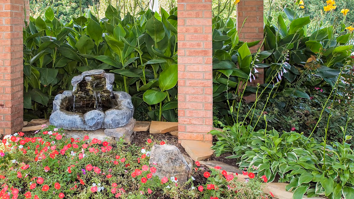 A small outdoor garden area with a stone water fountain surrounded by lush green plants and colorful flowers, framed by brick pillars.