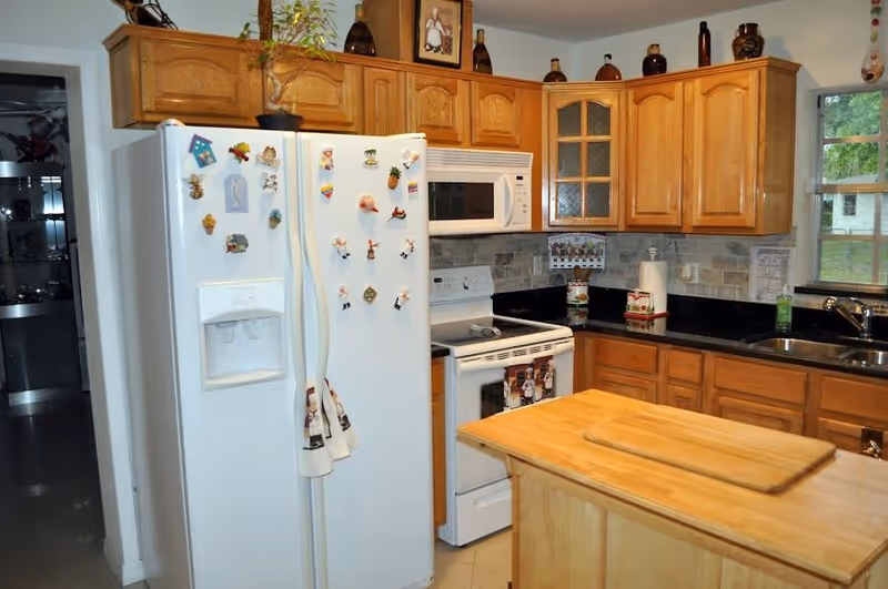 A kitchen with wooden cabinets, a white refrigerator decorated with magnets, a white stove with an oven, a microwave above the stove, black countertops, a double sink under a window, and a wooden island with a cutting board on top.