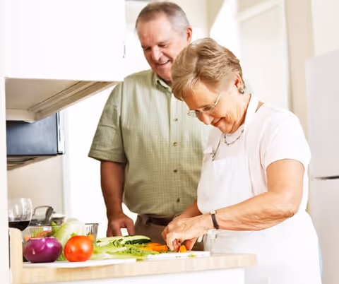 An elderly woman wearing glasses and a white apron is chopping vegetables on a kitchen counter while an elderly man in a green checkered shirt watches and smiles. Various fresh vegetables and a glass of red wine are visible on the counter.