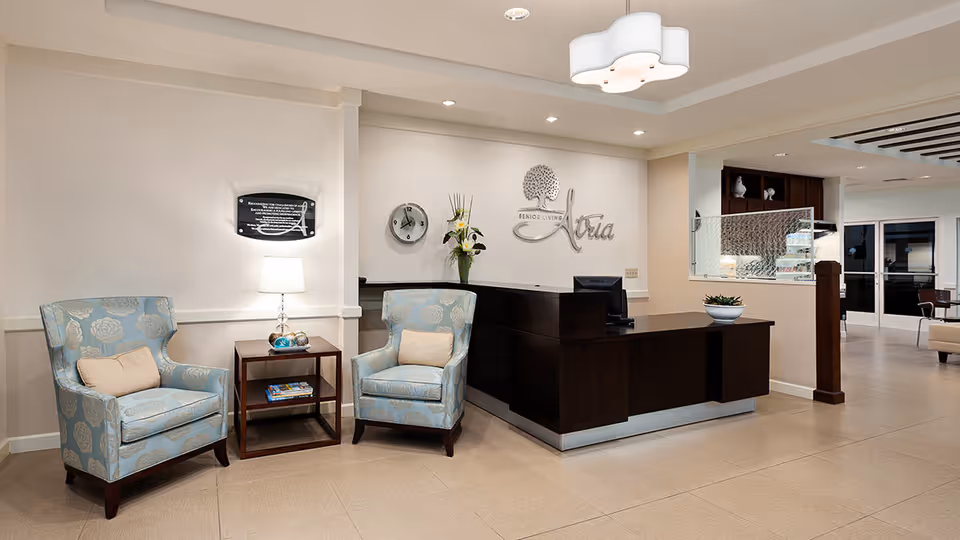 Reception area of Atria Palm Desert senior living facility featuring a dark wood reception desk with a computer, two light blue patterned armchairs with beige cushions, a small wooden side table with a lamp and books, a wall clock, and a decorative flower arrangement. The wall behind the desk displays the Atria Senior Living logo.