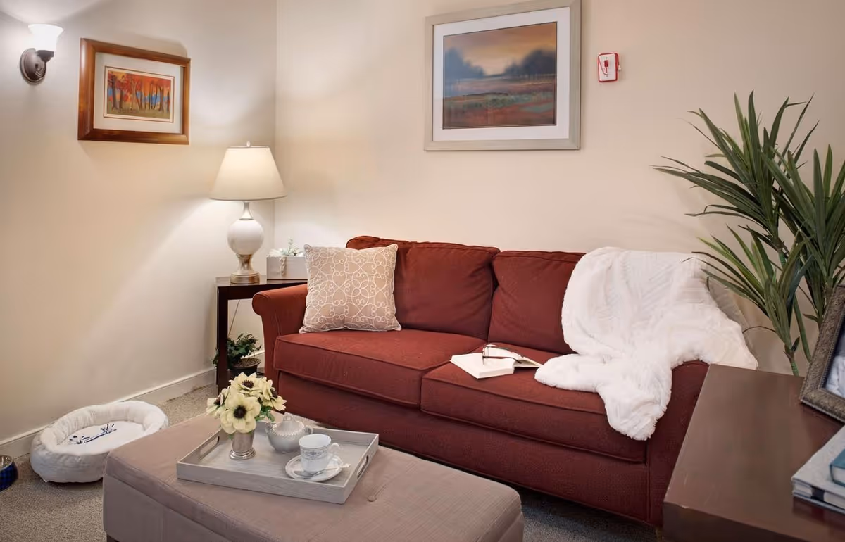 Cozy living room with a red sofa draped with a white throw, a lamp-lit side table, ottoman with a tray, and wall art.