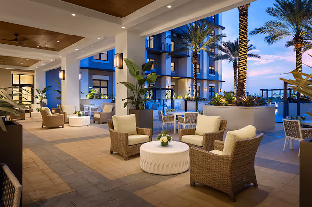 Outdoor covered patio area at dusk with wicker chairs and cushioned seating arranged around white circular tables, surrounded by potted plants and palm trees, with a modern building in the background.