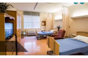 A nursing center patient room with two hospital beds, a reclining chair, TV cabinet, window and privacy curtains.