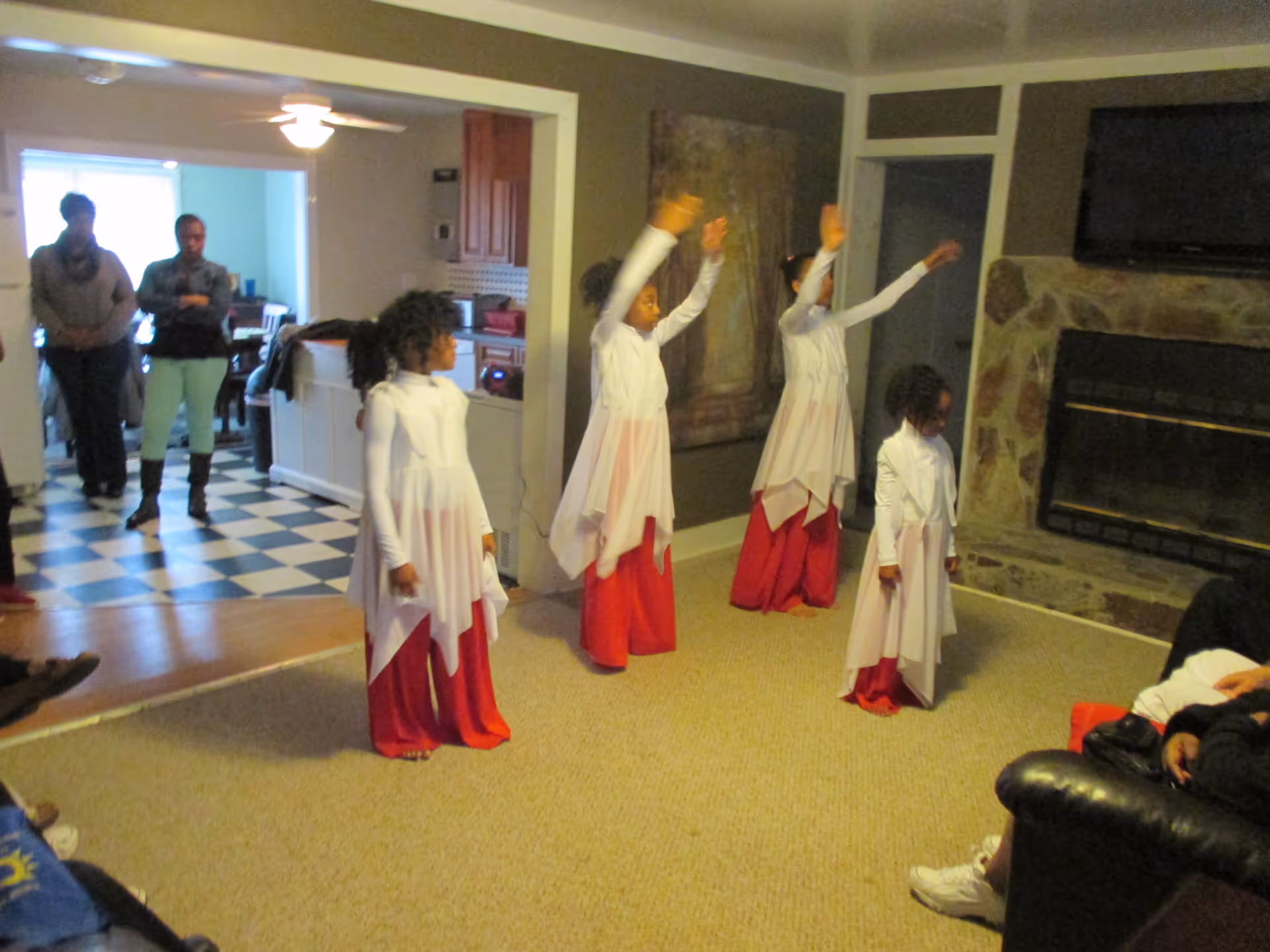 Four young girls dressed in white tops and red skirts are performing a dance in a living room with a stone fireplace and a mounted TV. Three adults stand in the adjacent kitchen area watching the performance. The room has beige carpet and neutral-colored walls.