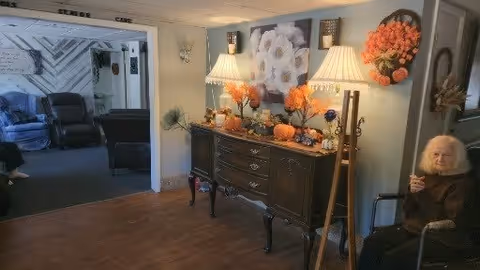 A cozy interior space featuring a dark wooden sideboard decorated with autumn-themed items including pumpkins, flowers, and two table lamps with white shades. Above the sideboard is a floral wall art. To the right, an elderly person with white hair is seated in a wheelchair. In the background, through an open doorway, a living room area with blue and black armchairs is visible.