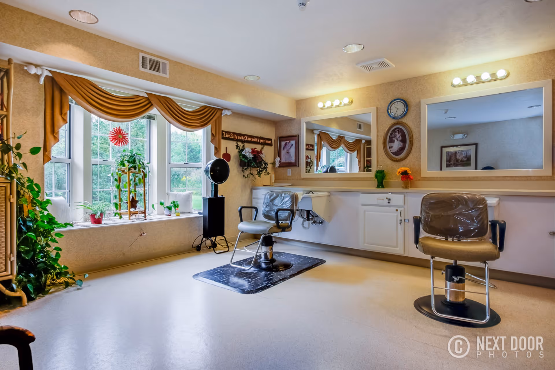 Interior of a salon area in a retirement village with two salon chairs in front of large mirrors mounted on the wall. The room has beige walls, a large window with mustard-colored drapes, several plants on the windowsill, and decorative wall art including a clock and framed pictures. The floor is light-colored and there is a hair dryer on a stand near the window.