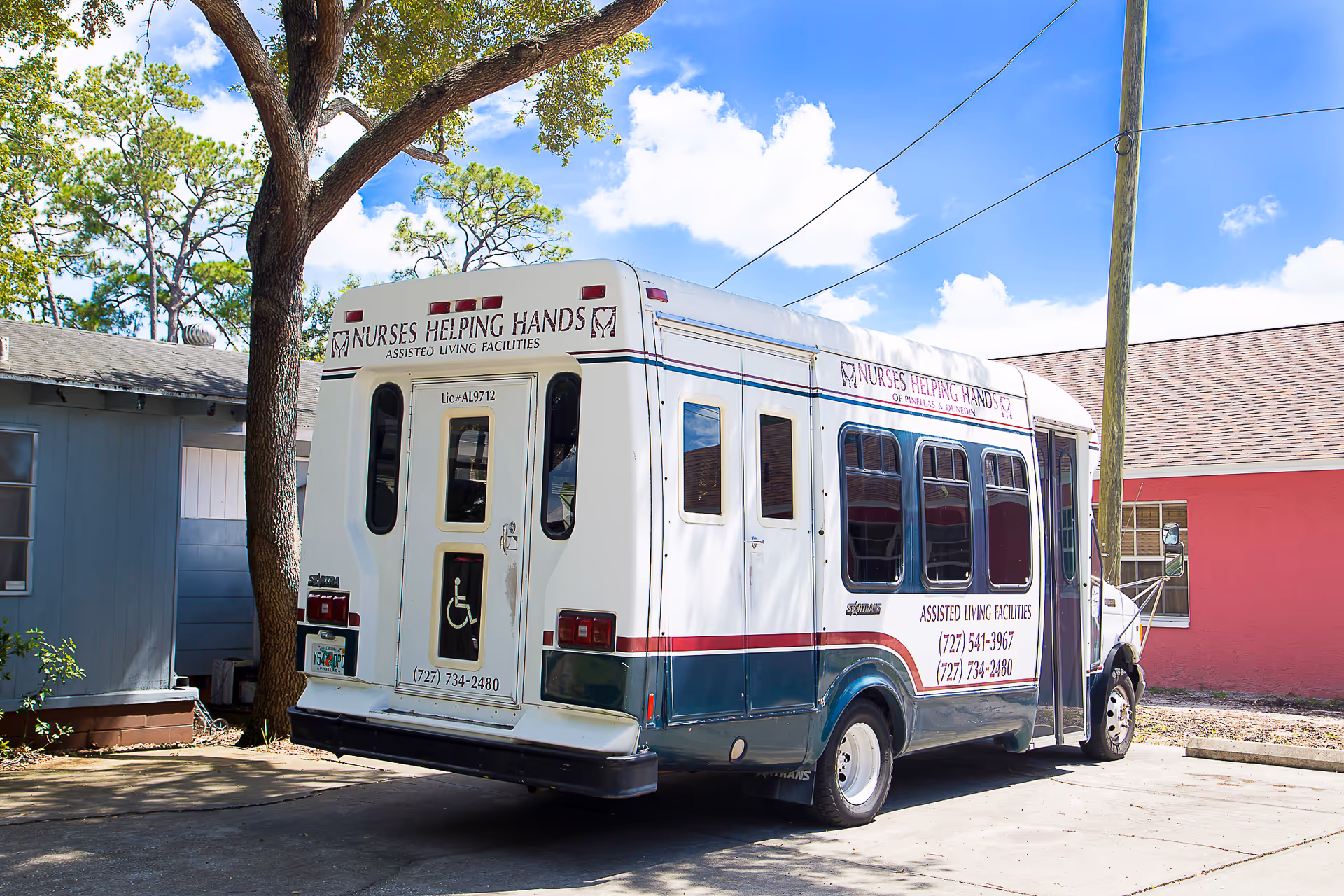 A white and green shuttle bus parked outside near a tree and buildings, with signage for Nurses Helping Hands Assisted Living Facilities, including phone numbers and a wheelchair accessibility symbol.