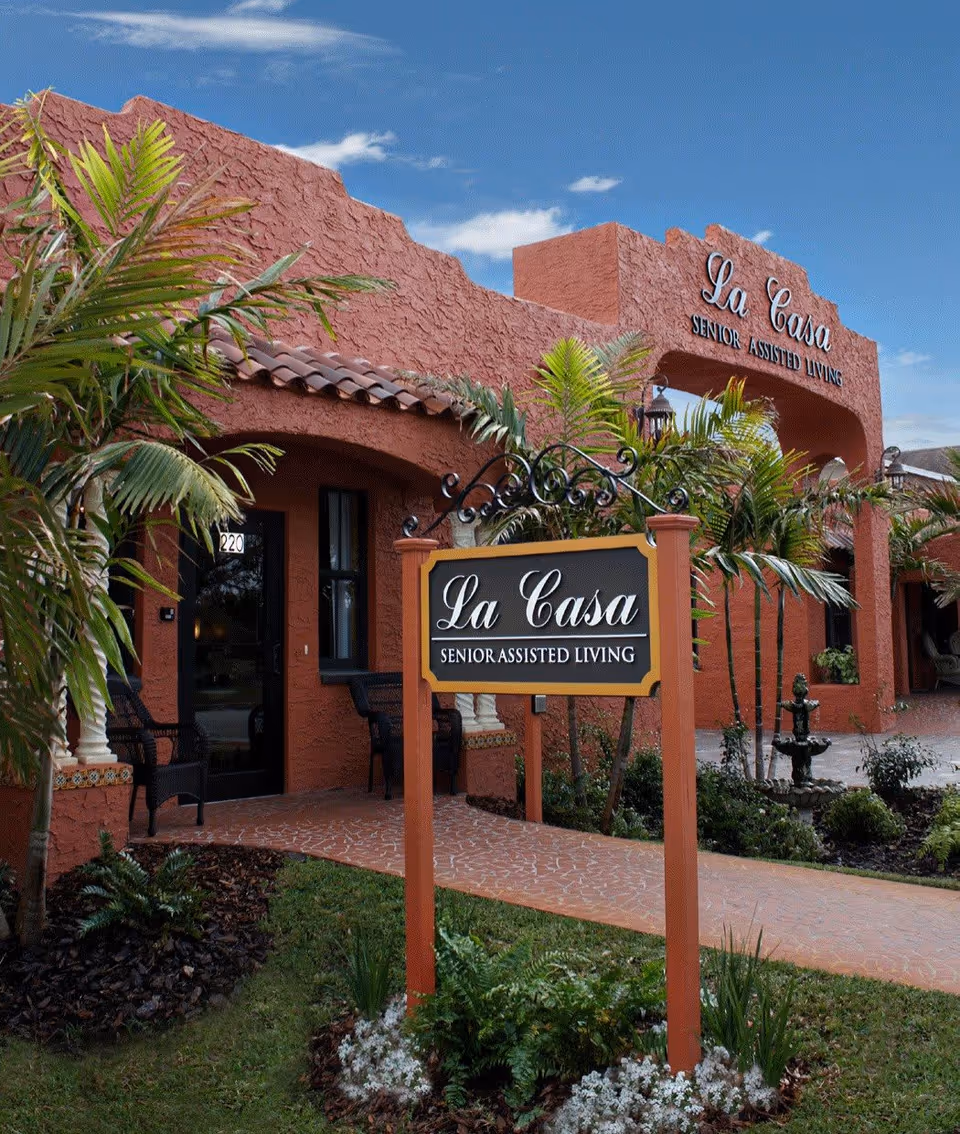 Exterior view of La Casa Senior Assisted Living building with a textured reddish facade, palm trees, a walkway, and a sign in front that reads 'La Casa Senior Assisted Living'.