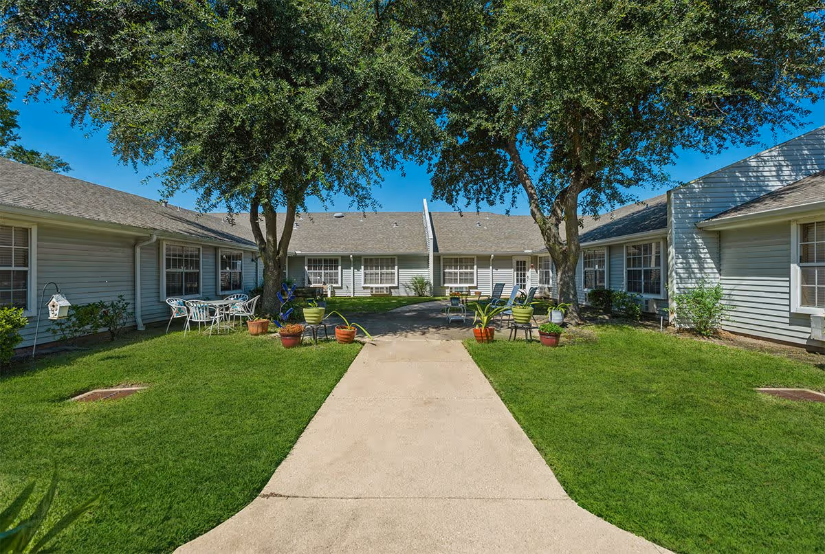 Outdoor courtyard area of a senior living facility with a concrete walkway leading to a seating area surrounded by green grass, potted plants, and two large trees. The courtyard is enclosed by single-story buildings with multiple windows under a clear blue sky.