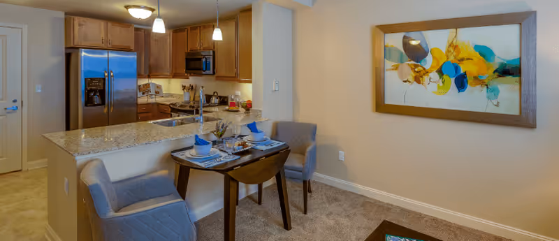 Interior view of a senior living facility apartment featuring a small dining area with a table set for two, two upholstered chairs, a kitchen with granite countertops, wooden cabinets, stainless steel refrigerator, microwave, and stove. A colorful abstract painting is hung on the wall beside the dining area.