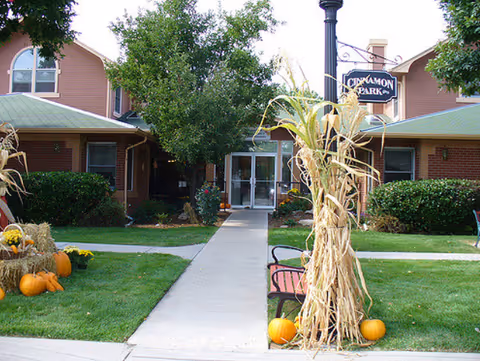 Front entrance of the Cinnamon Park building with a sidewalk, bench, lamp post, pumpkins and cornstalk decorations.