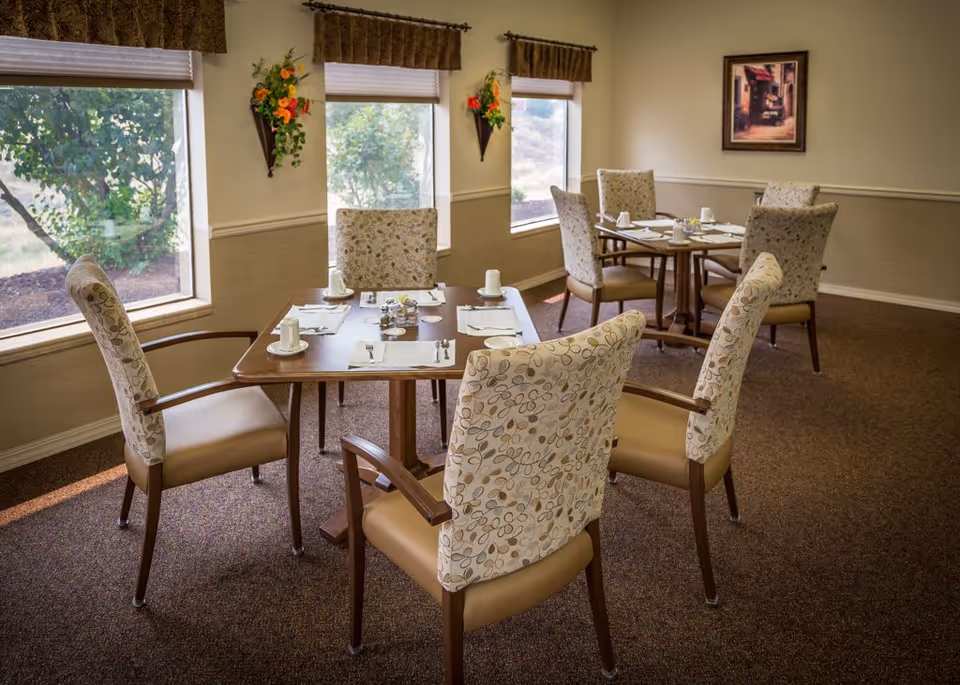 A dining room with two wooden tables, each set with white cups, saucers, and silverware. The chairs around the tables have patterned upholstery and wooden armrests. There are three large windows with brown valances letting in natural light, and wall-mounted flower arrangements between the windows. A framed picture hangs on the beige wall.