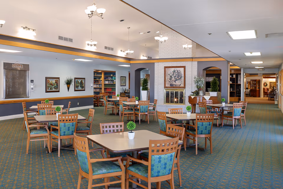 A spacious dining room in an assisted living facility with multiple square tables and wooden chairs with green patterned cushions. Each table has a small potted plant centerpiece. The room features a white brick fireplace with a floral painting above it, several chandeliers hanging from the ceiling, and a large mirror on one wall. There are bookshelves and framed artwork on the walls, and a carpeted floor with a green and orange pattern.