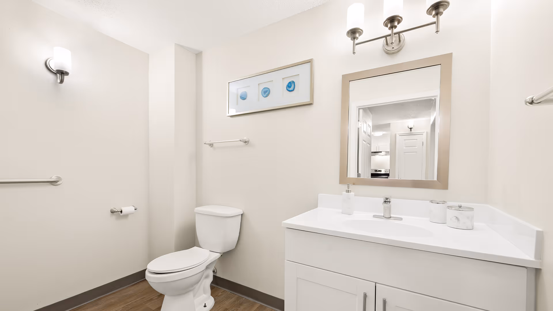 A clean and modern bathroom with a white toilet, white vanity with sink, a large mirror above the sink, a wall-mounted light fixture, a towel bar, and a framed artwork on the wall. The walls are painted light beige and the floor has wood-like flooring.