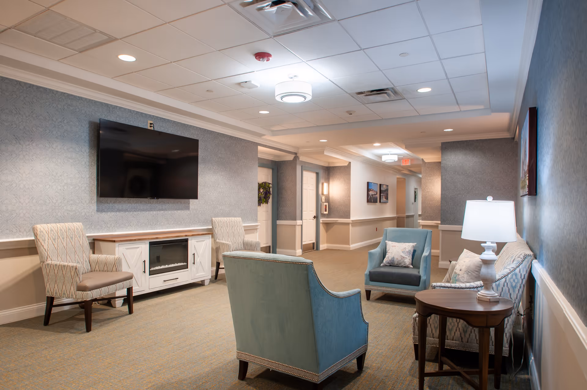 A cozy living room area in a senior living facility featuring a wall-mounted flat-screen TV above a white cabinet with an electric fireplace. The room has two patterned armchairs, a blue armchair, and a blue sofa with decorative pillows. A wooden side table with a white lamp is next to the sofa. The walls are painted in soft tones with a textured wallpaper, and the ceiling has recessed lighting and a central light fixture. The hallway beyond has framed pictures and doors leading to other rooms.