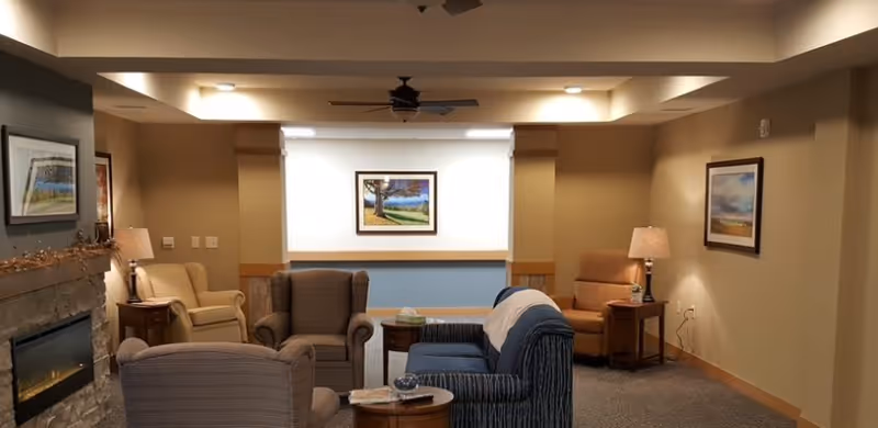 A cozy living room area in a senior living facility with several upholstered armchairs and a sofa arranged around small wooden tables. The room features a stone fireplace on the left, warm beige walls, framed landscape paintings, and soft lighting from table lamps. A ceiling fan is mounted above.