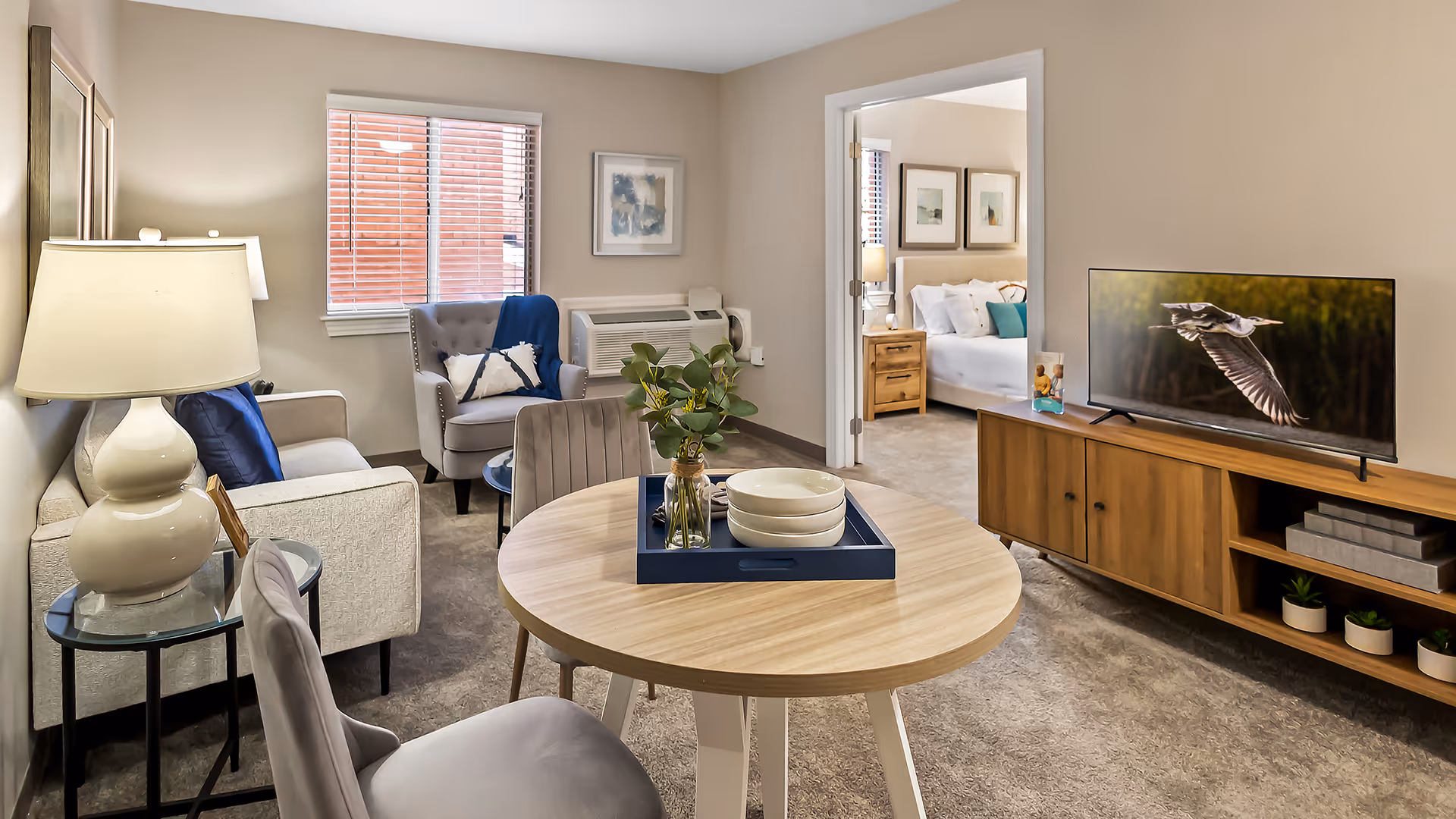 Furnished living area with round table, seating, TV on a wooden console, and an open doorway showing a bedroom.