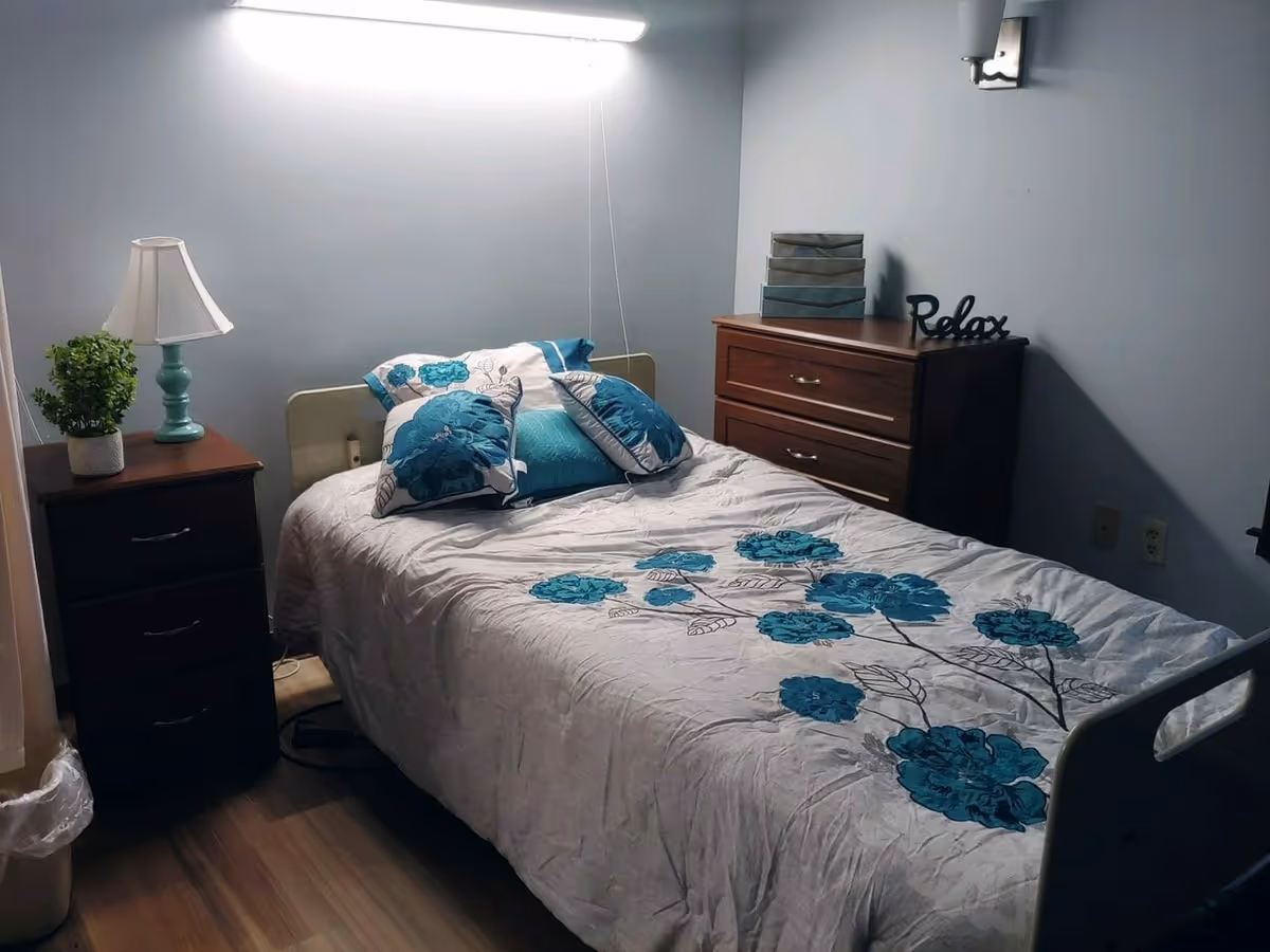 Small furnished bedroom with a single bed covered in teal floral bedding, a nightstand with a lamp and plant, and a wooden dresser.