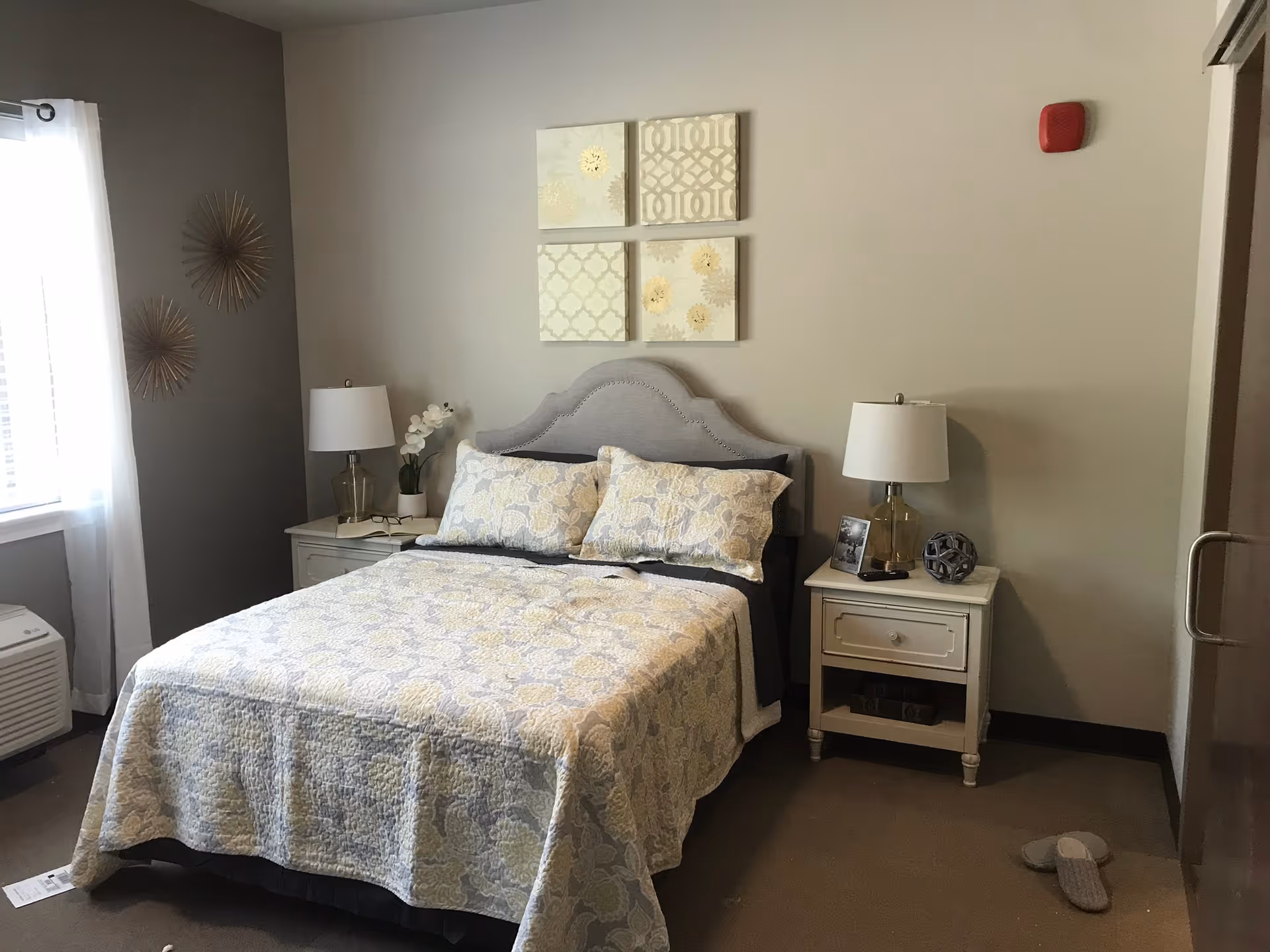 A neatly made bed with a patterned quilt and two pillows in a bedroom. There are two white nightstands on either side of the bed, each with a lamp. One nightstand has a small plant and the other has a framed photo and decorative item. Above the bed are four square wall art pieces with light patterns. A window with sheer white curtains is on the left side, and a pair of slippers is on the floor near the right nightstand.