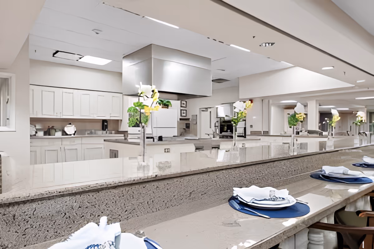 Interior view of a clean, modern kitchen area with white cabinets, stainless steel appliances, and a long counter set with plates, napkins, and cutlery. The counter has a textured front and is decorated with small flower arrangements. The ceiling has recessed lighting and a large stainless steel ventilation hood above the cooking area.