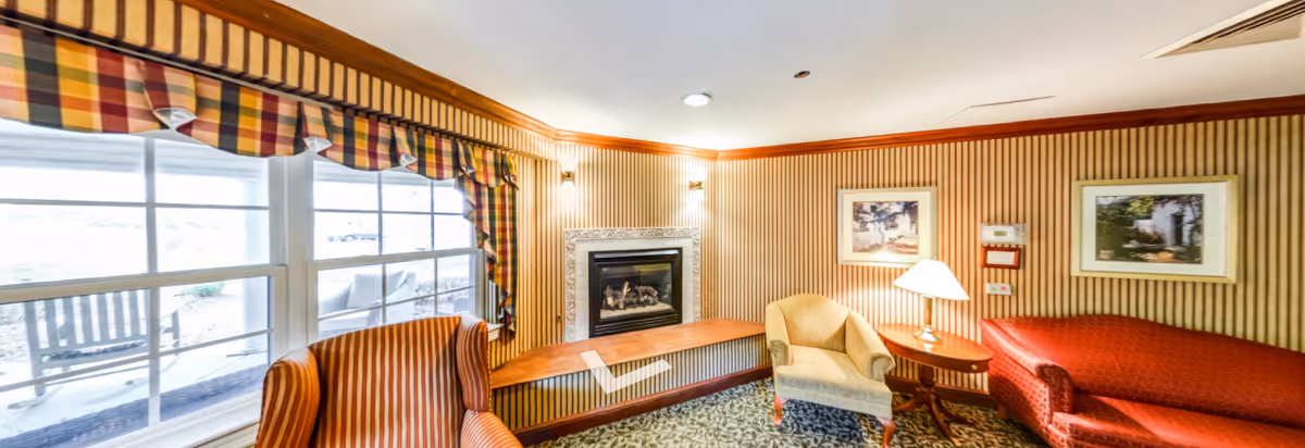 A cozy living room with striped wallpaper and a large window with plaid valances. The room features a fireplace with a marble surround, a red upholstered sofa, a beige armchair, a wooden side table with a lamp, and framed artwork on the walls. Outside the window, a porch with rocking chairs is visible.