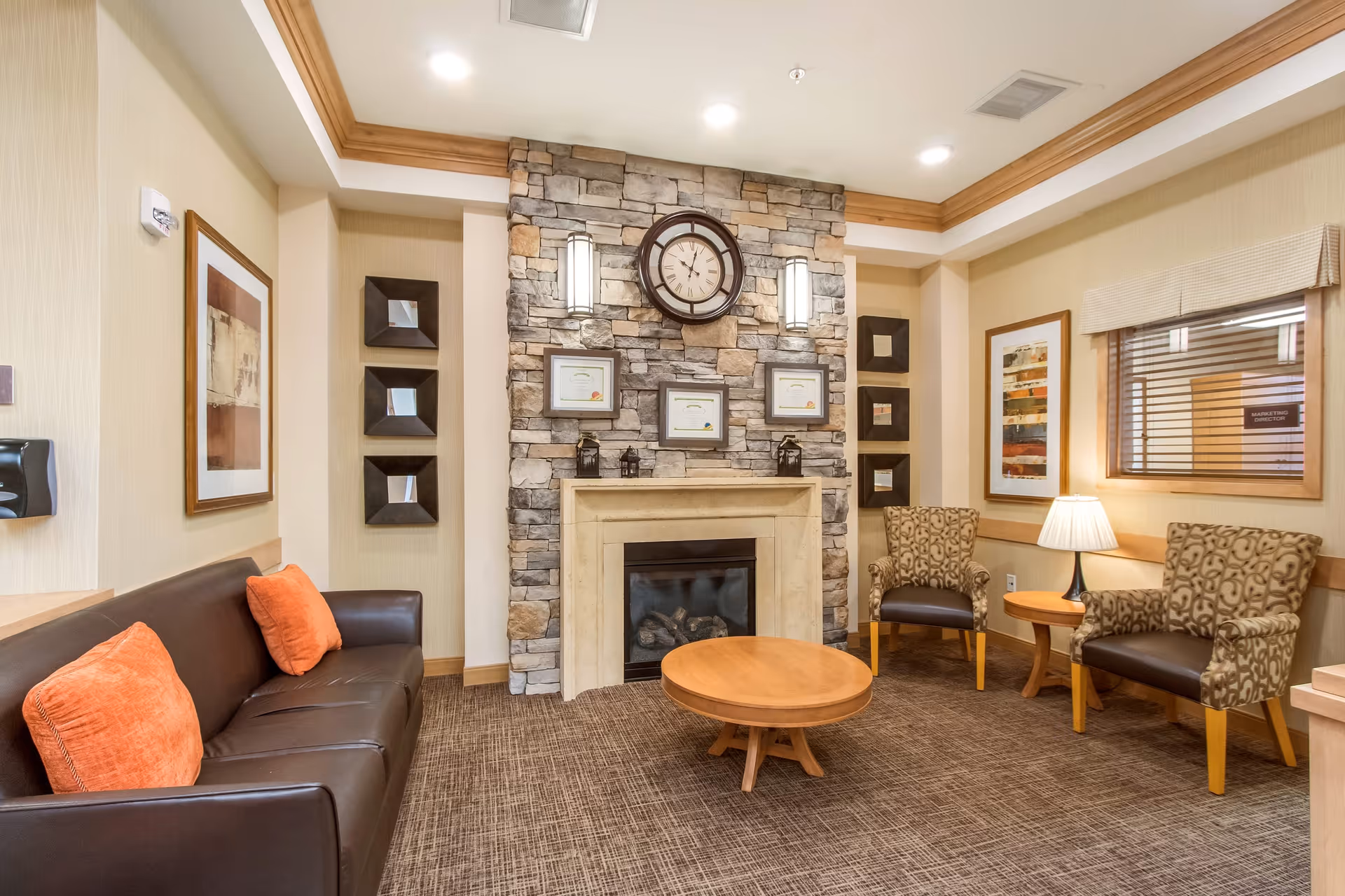 A cozy living room area with a stone fireplace in the center, above which hangs a large round clock. The fireplace mantel displays three framed certificates and two small lanterns. To the left is a dark brown leather sofa with two orange throw pillows. On the right side, there are two patterned armchairs with a small wooden side table and a lamp between them. The walls are decorated with framed artwork and small square mirrors. The room has beige walls, a carpeted floor, and recessed ceiling lights.