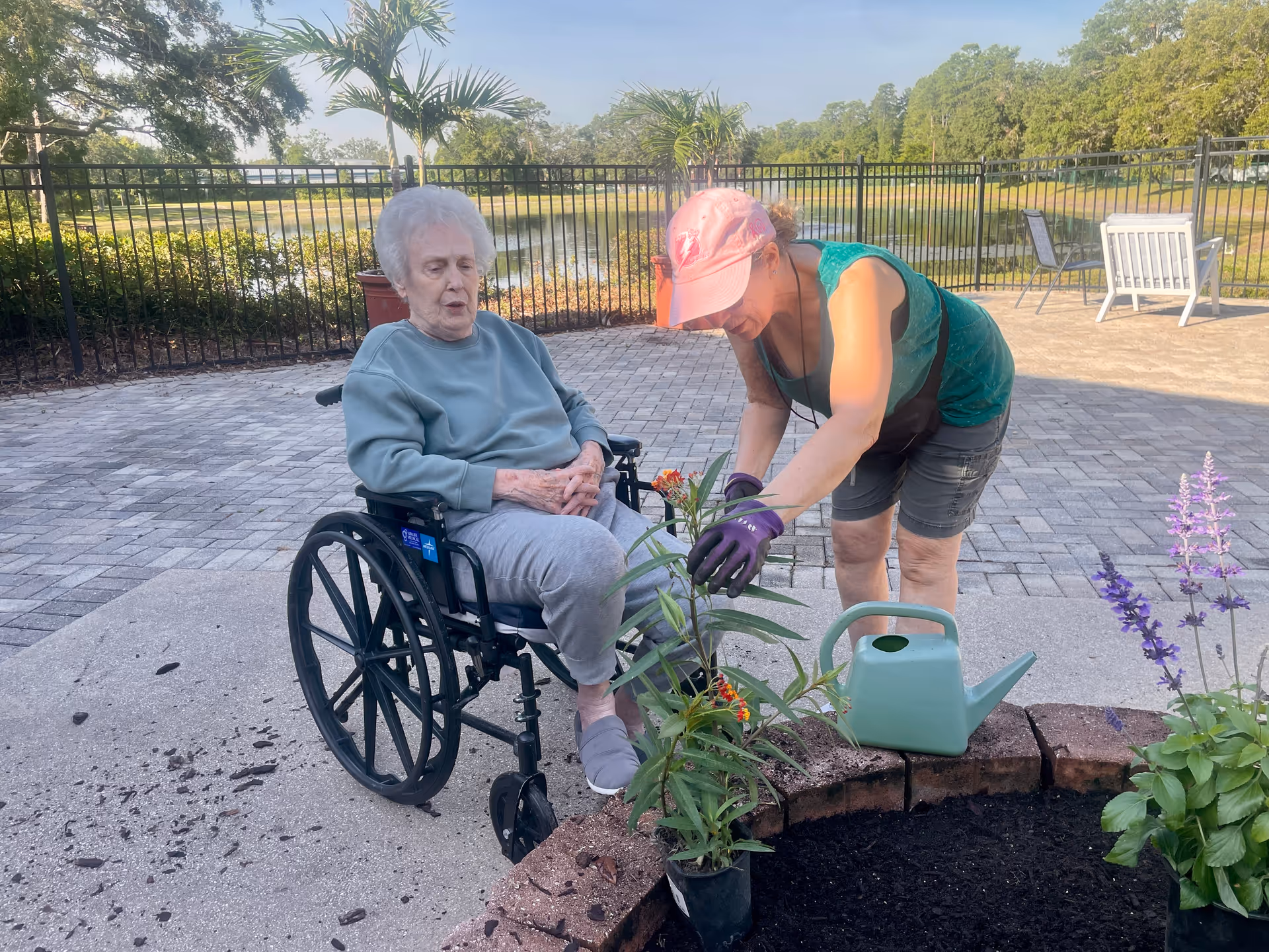 An elderly woman in a wheelchair is outdoors on a paved patio near a garden bed. A younger woman wearing a pink cap, green sleeveless top, and gloves is tending to plants in the garden bed, with a green watering can nearby. There is a black metal fence and trees in the background under a clear sky.