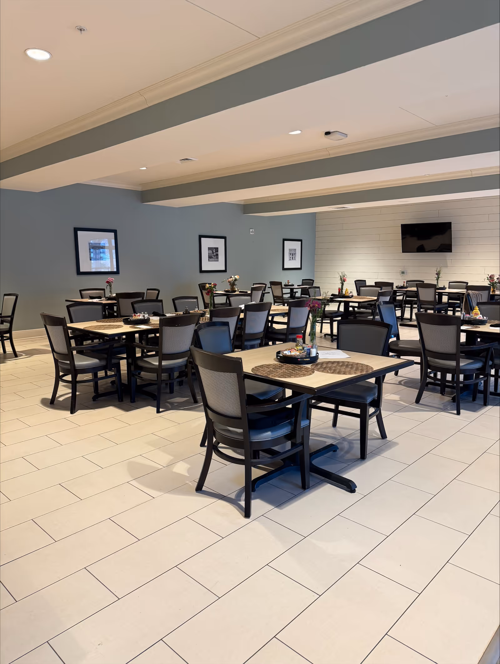 A spacious dining room with multiple square tables and chairs arranged neatly. Each table has placemats and some have small flower vases and condiment trays. The room has light-colored tiled flooring, a light blue wall with framed pictures, and a mounted flat-screen TV on a white shiplap wall.